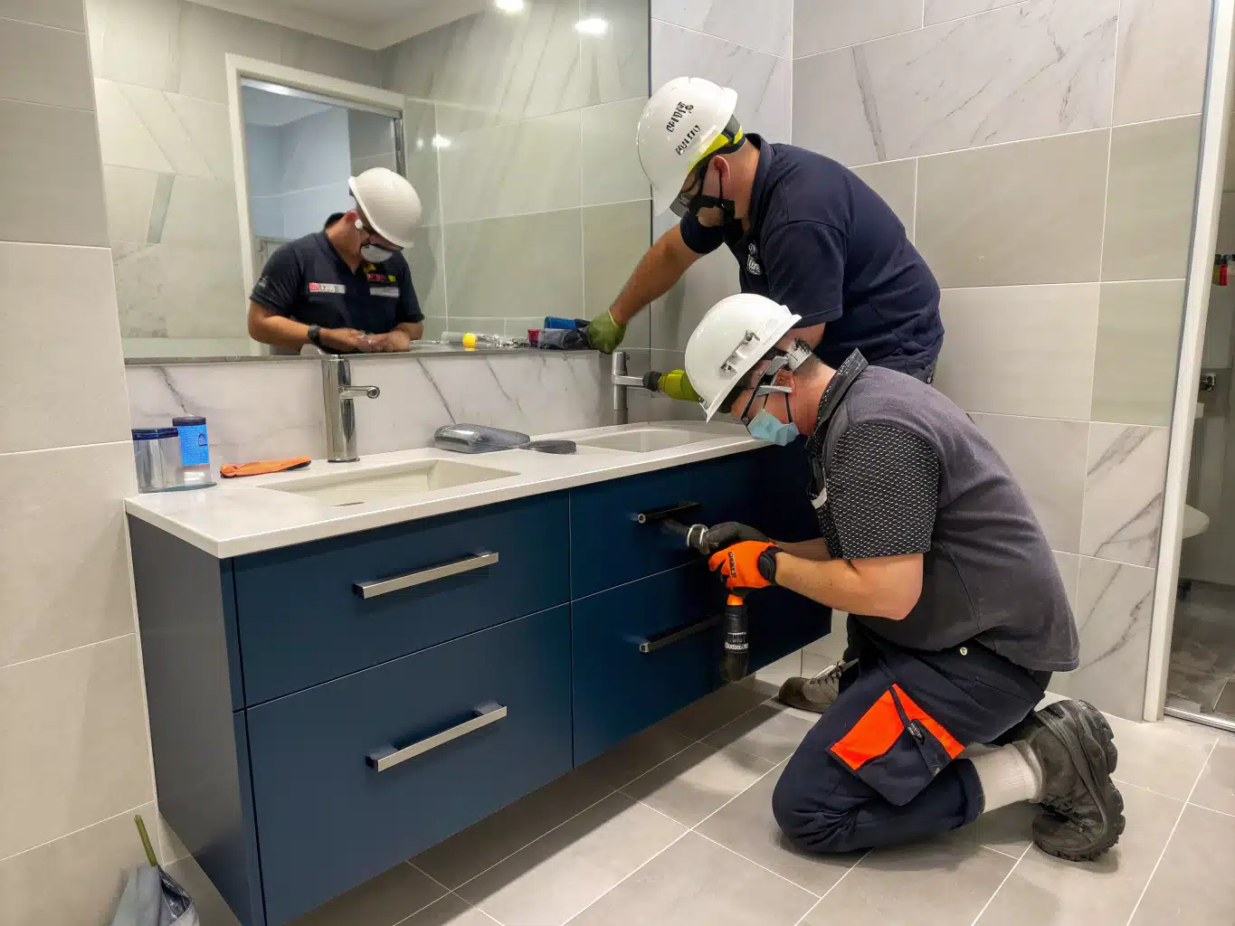 A dynamic scene of a small Sydney bathroom being transformed by skilled tradespeople, showcasing smart storage solutions and elegant fixtures. The workers, wearing safety helmets, gloves, and eyewear, are actively installing sleek cabinetry in deep indigo and midnight blue tones, complemented by light neutral tiles and charcoal accents. One worker is carefully measuring and cutting a modern vanity while another is organizing tools and materials, emphasizing efficiency in a compact space. The atmosphere is vibrant and collaborative, capturing the essence of innovation and craftsmanship in an authentic working environment.